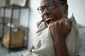 Happy energetic young dark skinned man in eyewear sitting in bedroom squeezing pillow, opening mouth excitedly, having joyful facial expression. Bedtime, comfort, leisure, energy and vitality