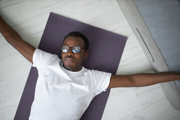 High angle view of peaceful tired young African male in glasses and tee lying on mat with arms...