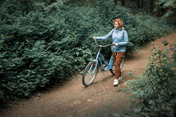 A blonde woman in a blue jacket walks through the forest with a blue bicycle
