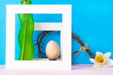 Still life green vase, egg and crown of thorns in a white frame on a blue background. On the crown of thorns is a narcissus flower.