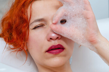 Fototapeta premium Portrait of a young beautiful woman in bathroom, looking through foam between fingers