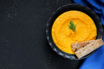 Carrot and pumpkin puree soup with bread croutons garnished with dill in a black plate on a dark background with a chalk effect. Blue napkin. Copy space. Top view