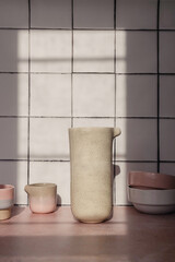 Handmade empty tableware such as ceramic jars, mugs and dishes composition on a white kitchen tile background on a sunny day with shadows. Pink, blue and beige color glaze on clay. Creative utensils.