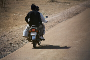 Two people on moped with milk can on the luggage rack seen from behind on a dusty country road in...