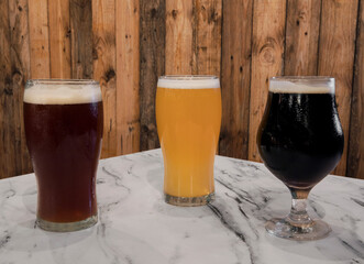 Brew. Closeup view of three glasses with different kinds of beers, black, red and golden, on the pub white marble table with a wooden background.