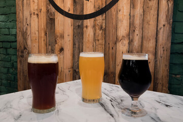 Brew. Closeup view of three glasses with different kinds of beers, black, red and golden, on the pub white marble table with a wooden background.