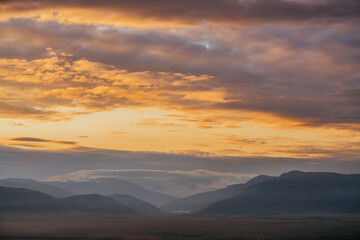 Scenic dawn mountain landscape with golden low clouds in valley among mountains silhouettes under cloudy sky. Vivid sunset or sunrise scenery with low clouds in mountain valley in illuminating color.