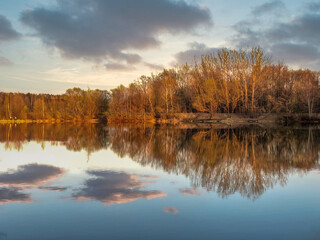 Golden hour sunlight at forest lake with still water surface reflecting clousd and bare trees on distant shore in spring evening