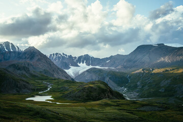 Dramatic landscape with mountain lake and big glacier under cloudy sky. Spot of sunlight on mountains in overcast weather. Atmospheric mountain scenery with cloudiness above great mountain range.