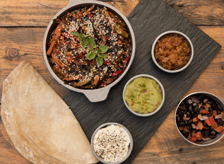 Gourmet vegetarian food. Spicy flavors. Overhead closeup view of Mexican fajitas with bowls containing spicy vegetables, sauces and dips in a black plate on the wooden table.