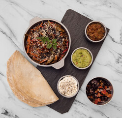 Gourmet food presentation. Vegetarian cuisine. Overhead view of burritos and bowls with spicy cooked vegetables, sauces and dips in a black plate on the white marble table.
