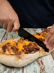 Gastronomy. Rustic flavors. Closeup view of a male caucasian chef slicing a cheddar, mozzarella cheese, bacon and chicken pizza with a black knife on the restaurant wooden table. 