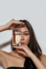 Young asian woman hiding face behind glass with water - obrazy, fototapety, plakaty