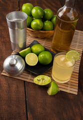 Brazilian drink caipirinha, glass of caipirinha, bottle of cachaça, cocktail shaker and lemons on wooden mat and rustic wood light background, selective focus.