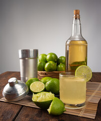 Brazilian drink caipirinha, glass of caipirinha, bottle of cachaça, cocktail shaker and lemons on wooden mat and rustic wood light background, selective focus.