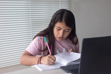 Little girl writing on a notebook. Doing homework at home with the laptop.