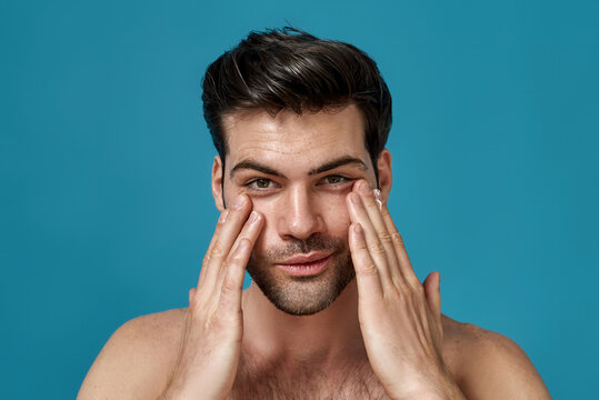 Beauty portrait of handsome man looking at camera with a smile while applying moisturizing cream on his face isolated over blue background