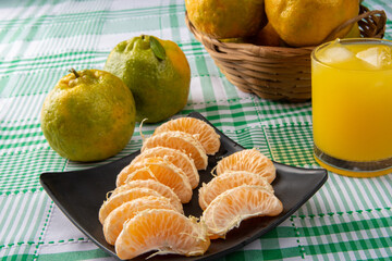 Orange ponkan, orange ponkan slices on a black plate, plus some oranges and a glass of juice on a checkered tablecloth on a light background, selective focus.