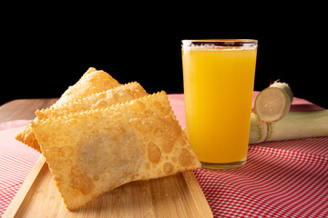 Brazilian fried pastries, a glass of sugarcane juice and canes positioned on a checkered tablecloth, black background, selective focus.