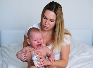 A young mother feeds her baby from a bottle with milk. The baby is crying, his teeth are teething, he wants to sleep or he has a stomach ache.