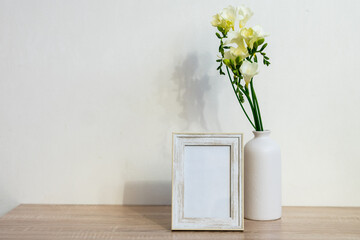 Portrait white picture frame mockup on wooden table. Modern ceramic vase with yelow freesia flowers . White wall background. Scandinavian interior. 