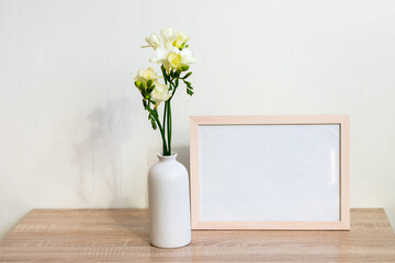 Portrait white picture frame mockup on wooden table. Modern ceramic vase with yelow freesia flowers . White wall background. Scandinavian interior. 