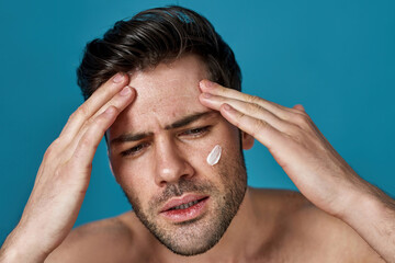 Beauty portrait of serious confident guy with brown hair looking aside while applying cream on his face, posing isolated over blue background