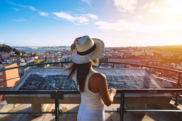 Young carefree woman tourist in white hat looking at Lisbon city from viewpoint at sunset. Tourism in Europe. Travel, vacation and holidays concept.  - obrazy, fototapety, plakaty