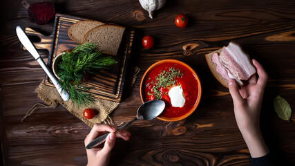 Hands holding a spoon and piece of bacon sandwich. Traditional Ukrainian borsht, red vegetable and meat soup or borscht with smetana on wooden background. Slavic dish with cabbage, beets, tomatoes
