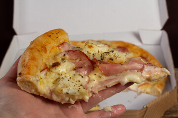 Hand holding a slice of pizza of pork loin with cream cheese and pineapple or lombo ao creme com abacaxi; pizza box in the background