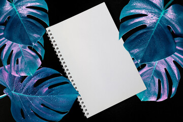 Art image of Monstera leaves and a white notepad on a black background. Blue leaves in the cyberpunk style. Copy space