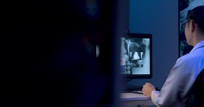 Asian male doctor is making video call conference on monitors showing x-ray film. Alone man medical assistant is consulting telehealth with senior patient at online chat at late night. Dolly shot.