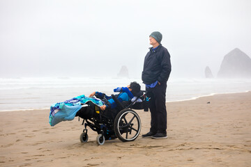 Obraz premium Father with disabled son in wheelchair on cold foggy beach