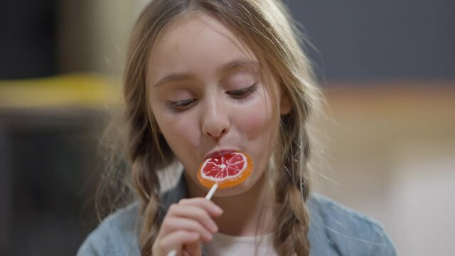 Headshot of joyful Caucasian girl with ponytails licking lollypop looking at camera and smiling. Close-up portrait of happy relaxed cheerful schoolgirl posing with sweet dessert in classroom in school