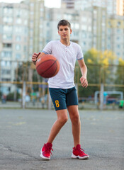 A cute young boy plays basketball on the street playground in summer. Teenager in a white t-shirt with orange basketball ball outside. Hobby, active lifestyle, sports activity for kids.