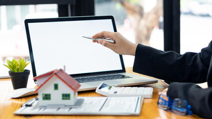 A real estate agent introduces clients to a home layout with contract documents and a laptop and dollar bill on the table. Customers interested in purchasing a home sign their signature to complete a 
