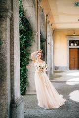 Bride in a beautiful dress with a bouquet of pink flowers stands in the vaulted hall with her hand on her forehead. Lake Como, Italy