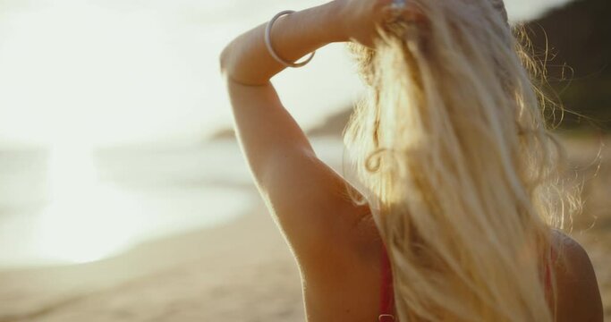 Beautiful young woman relaxing on the beach at sunset golden hour, long blond hair and red bikini, looking out at the waves, summer beach lifestyle