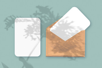 Mockup with an overlay of plant shadows on envelope with two sheets of textured white paper on a green table background. Horizontal orientation