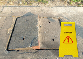 Grade separation of old concrete damaged sewer or drain pipe on sidewalk or footpath. Social problem concept. "DANGER" sign and symbol and exclamation mark icon on the yellow board.
