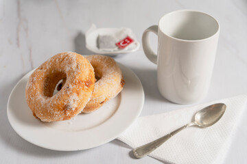 Pair of donuts with sugar and filled with sweet on a white plate with a white cup on a marble table.