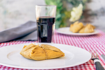 Tamale served on a white plate on a traditional table next to a glass of red wine. A typical sandwich or Latin American meal of cornmeal and meat. Traditional Andean food. Traditional food concept