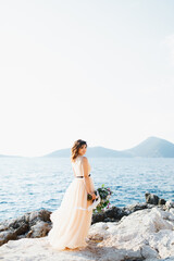 Fototapeta na wymiar Smiling bride in a pastel wedding dress stands on a rock above the sea with a bouquet of flowers in her hands
