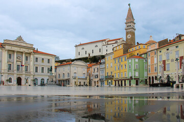 Fototapeta premium The main square of Piran with colorful houses, shops, cafes, a monument and a town hall with reflections in a puddle.