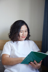 Young Asian Pregnant Woman reading book on the bed at home while holding her belly 