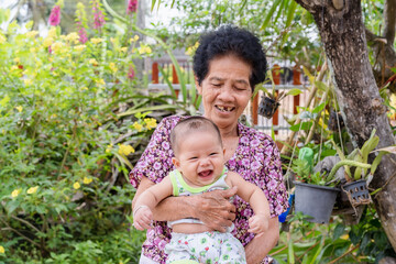 Happy big laughing child boy and senior woman holding adorable baby boy in flowery garden. Grandmother holding newborn baby grandson outdoors with nature background. Family and healthcare concept