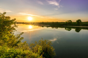 Fototapeta na wymiar Scenic view at beautiful summer river sunset with reflection on water with green bushes, grass, golden sun rays, calm water ,deep blue cloudy sky and glow on a background, spring evening landscape