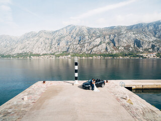 Fototapeta na wymiar Hugging newlyweds stand near a black convertible on the pier against the backdrop of the sea and mountains