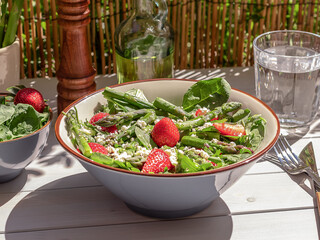 Fresh strawberry, green asparagus, feta cheese, baby spinach, and arugula salad served in a bowl with sparkling water. natural summer light and trendy strong reflection. Selective Focus.