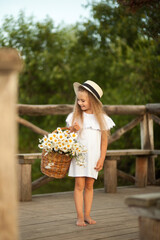 Little girl in a hat and white dress stands outdoors barefoot with a basket of flowers